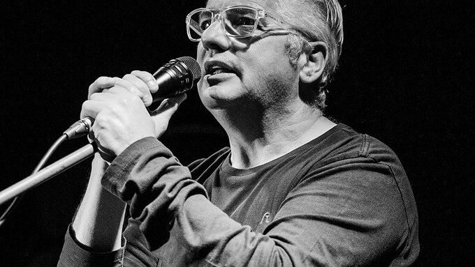 A black and white picture of JJ Gilmour - he has white hair, wears glasses and is singing into a microphone.