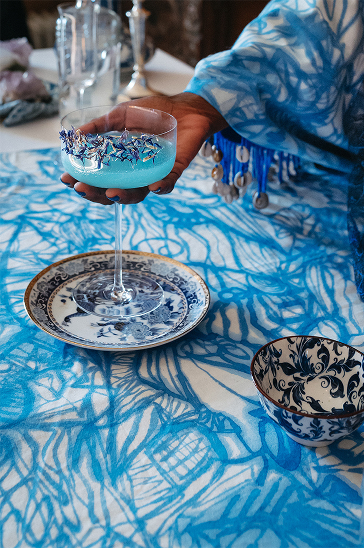 A photo of Sekai's hand placing a blue drink in an ornate glass on to bright blue streaked table cloth.
