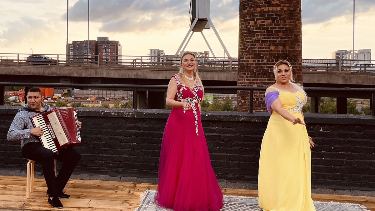 A photo of three performers on a rooftop, on the left a man plays an accordion to the right two women appear to dance