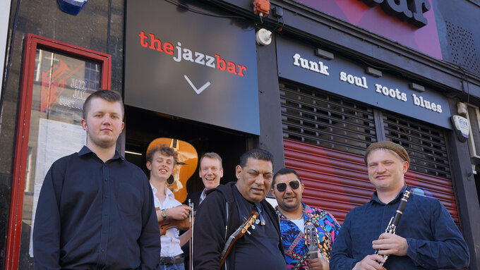 A group of six men of various ages stood in front of a jazz bar. They hold various instruments.