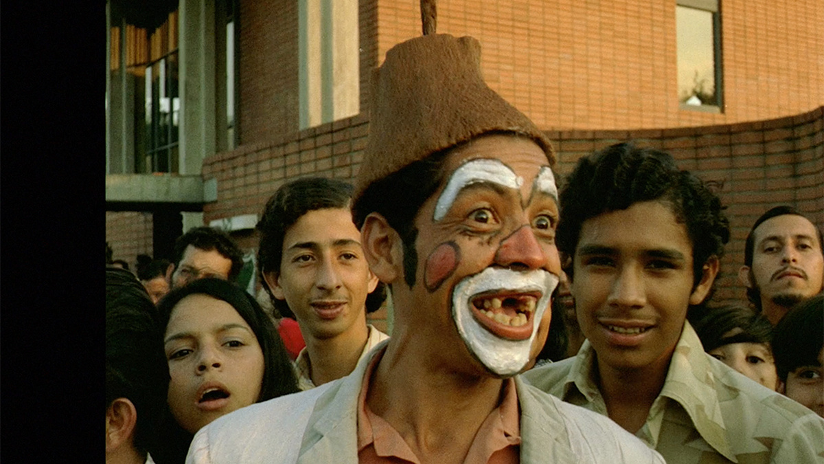 A man wearing a hat, a peach-colour shirt and jacket is in a crowd of people. He is missing teeth and has clown makeup.