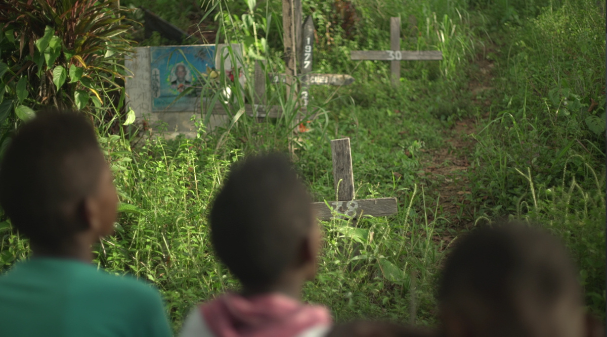 Three Afro-Colombian boys are pictured from behind as they look at a grassy cemetery with several wooden crosses.