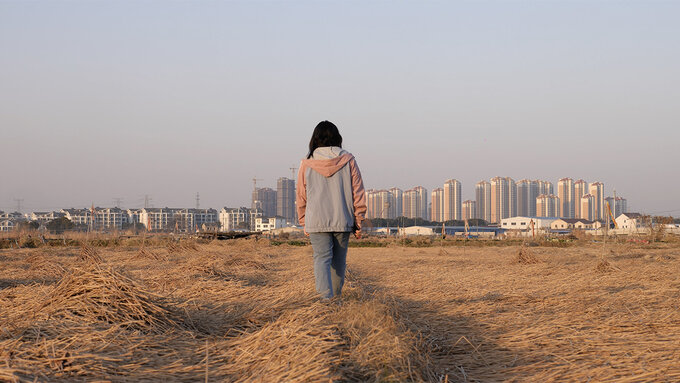 A woman walks away from the viewer through a field of straw, in the background is a clear smoggy sky and city scape