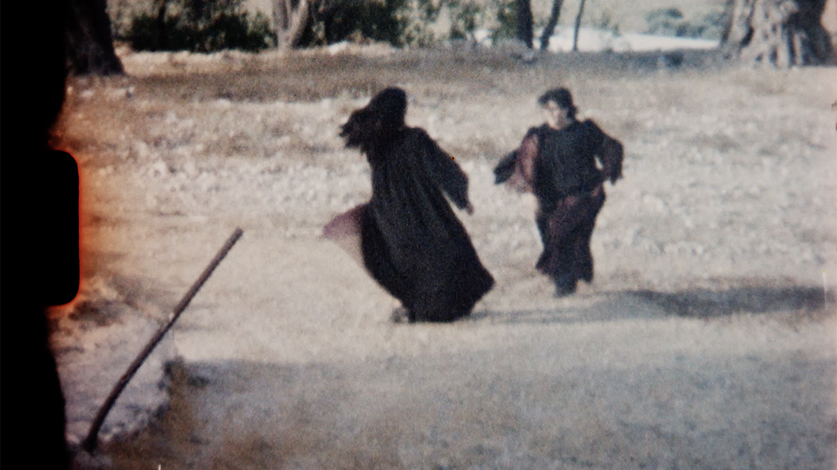 A film still of two figures with long hair in long flowing black robes, you can see a portion of the film's edge.