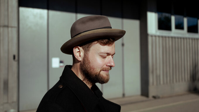 A photograph of John Lemke, he is looking down, and wears a beard and brown hat.