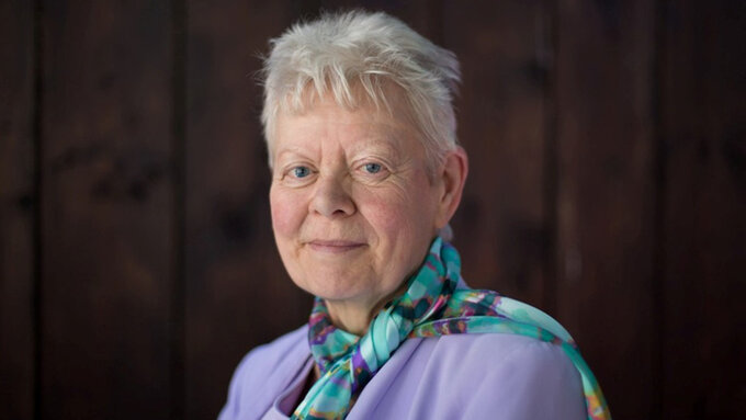 A photograph of A C Clarke smiling at the camera, she has short white hair and wears a silk scarf and a lavender jacket