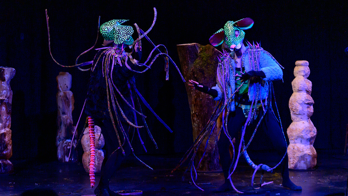 Two figures in sci-fi rat costumes face each other mid-dance they’re surrounded by surreal objects on a stage