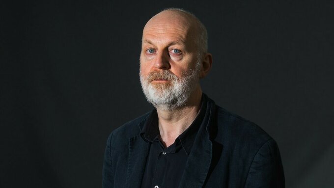 A photograph of Don Paterson, he has a white beard and is standing against a dark background.