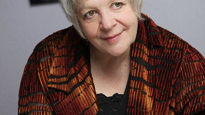 A photograph of Liz Lochhead, she has white hair and is wearing a brightly patterned orange blazer.