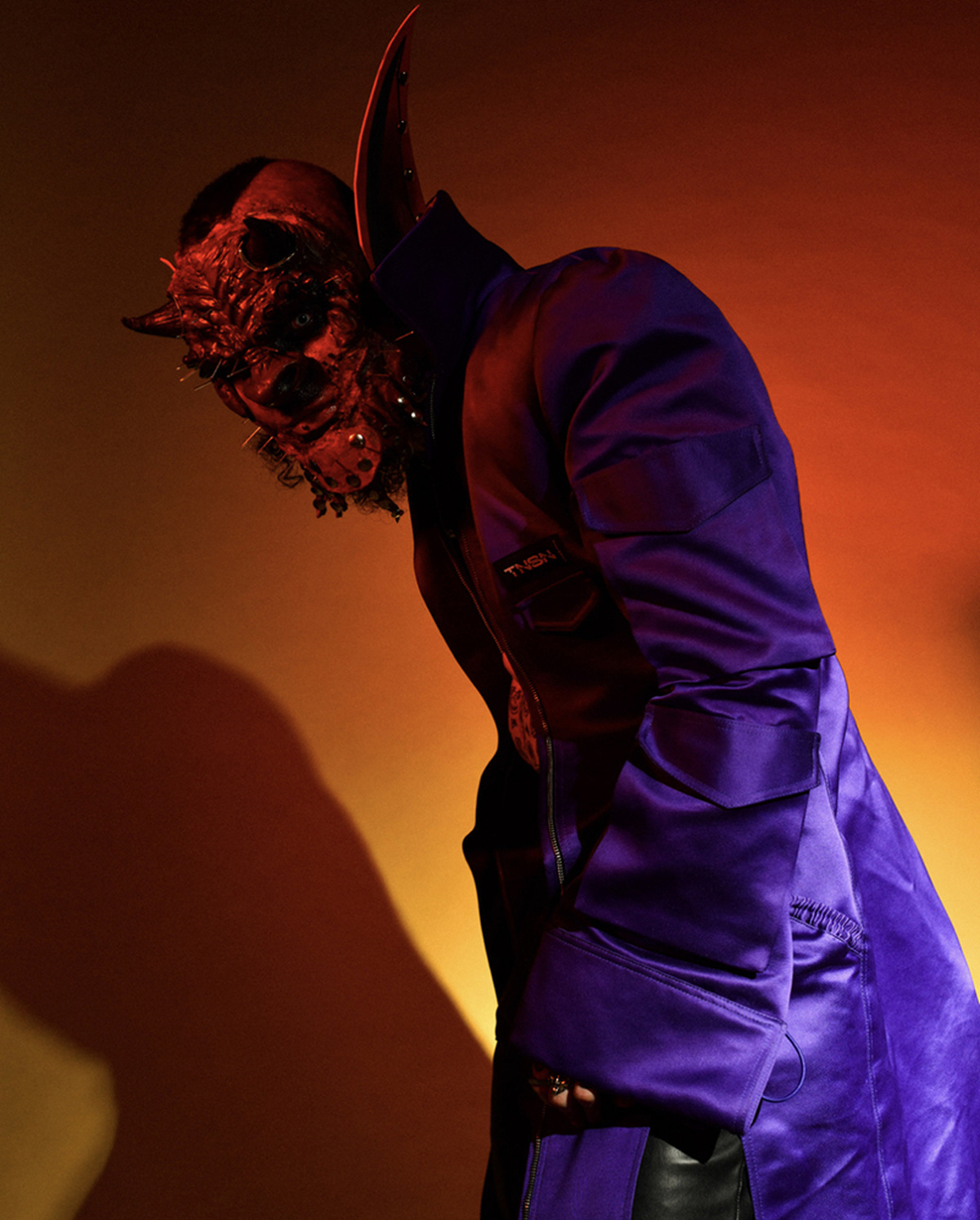 A low angle photo of a man dressed as a devil wearing a long overcoat, lit with dark orange light
