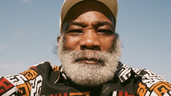 A picture of Dennis Bovell, he has a grey beard and wears a colourful patterned jumper and baseball cap.