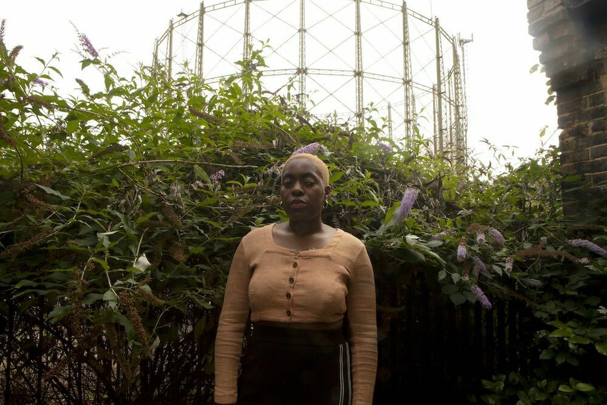 Onyeka stands in front of a green bush with purple flowers, wearing hoop earrings, a brown shirt and a black tracksuit.