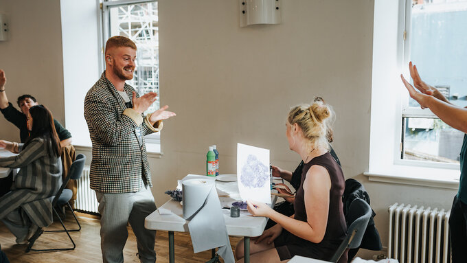 Busy room with many people making collages. Jamie Rea is talking to a participant in a workshop in BSL.