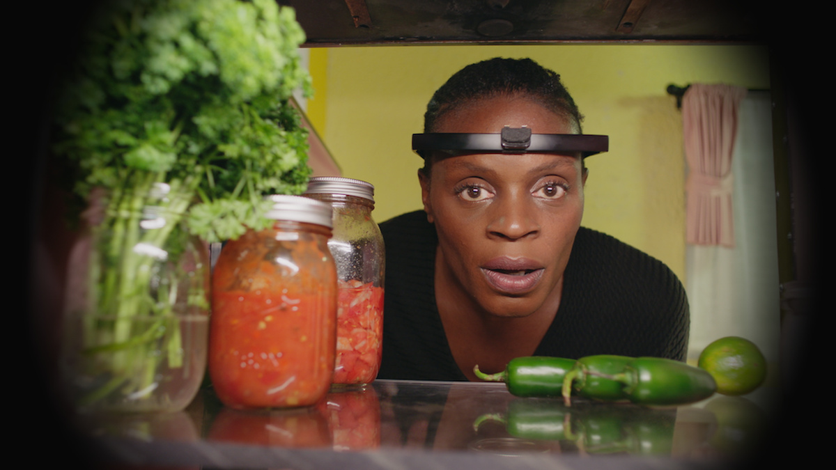 A woman appears surprised as she looks into the back of a refrigerator, which is stocked with vegetables and jars.