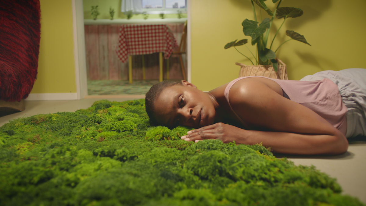 A woman lies on the floor of a home, her head resting on a mossy rug, and looks off into the middle distance.