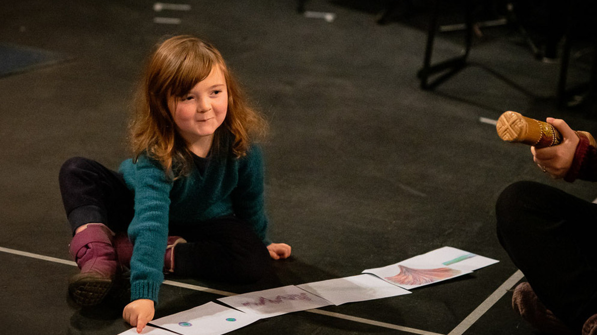 A girl plays in a GIObabies workshop, pointing at pictures on the floor while somebody plays percussion instrument.