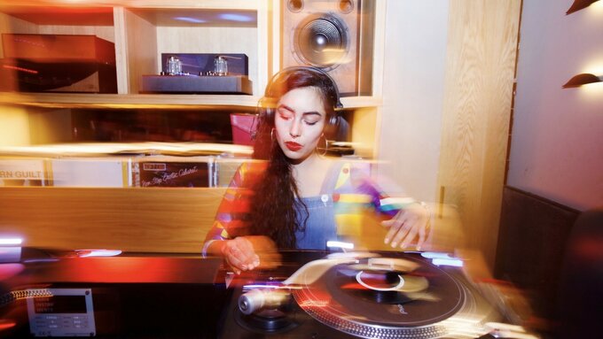 Cami is positioned at a DJ deck, they have long hair and wear red lipstick.