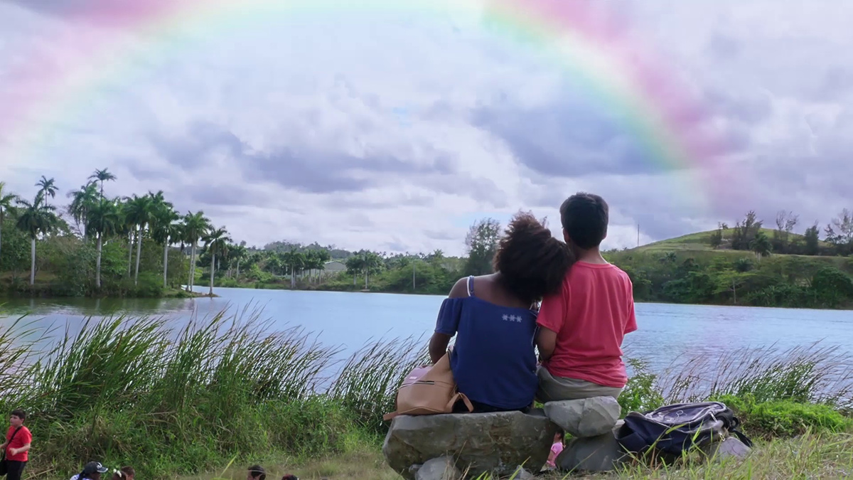 Sitting on a rock by a lake looking at a rainbow, a child puts their head on the shoulder of another child