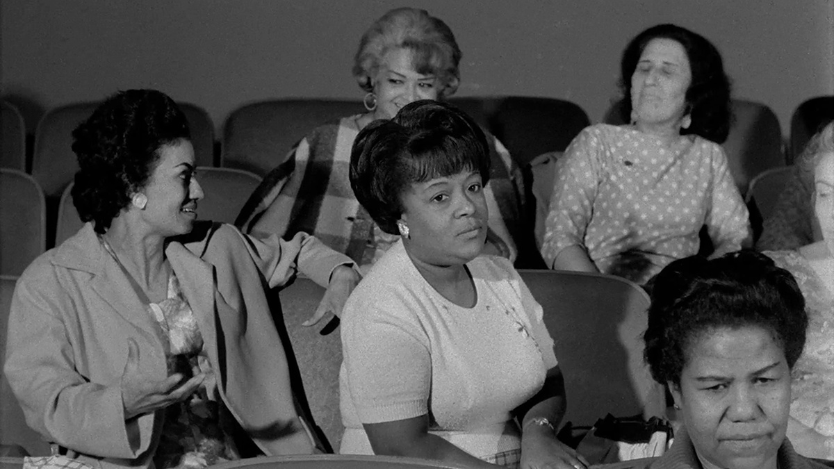 A group of people sit on seats in rows, chatting. The image is black and white and not from the present day.