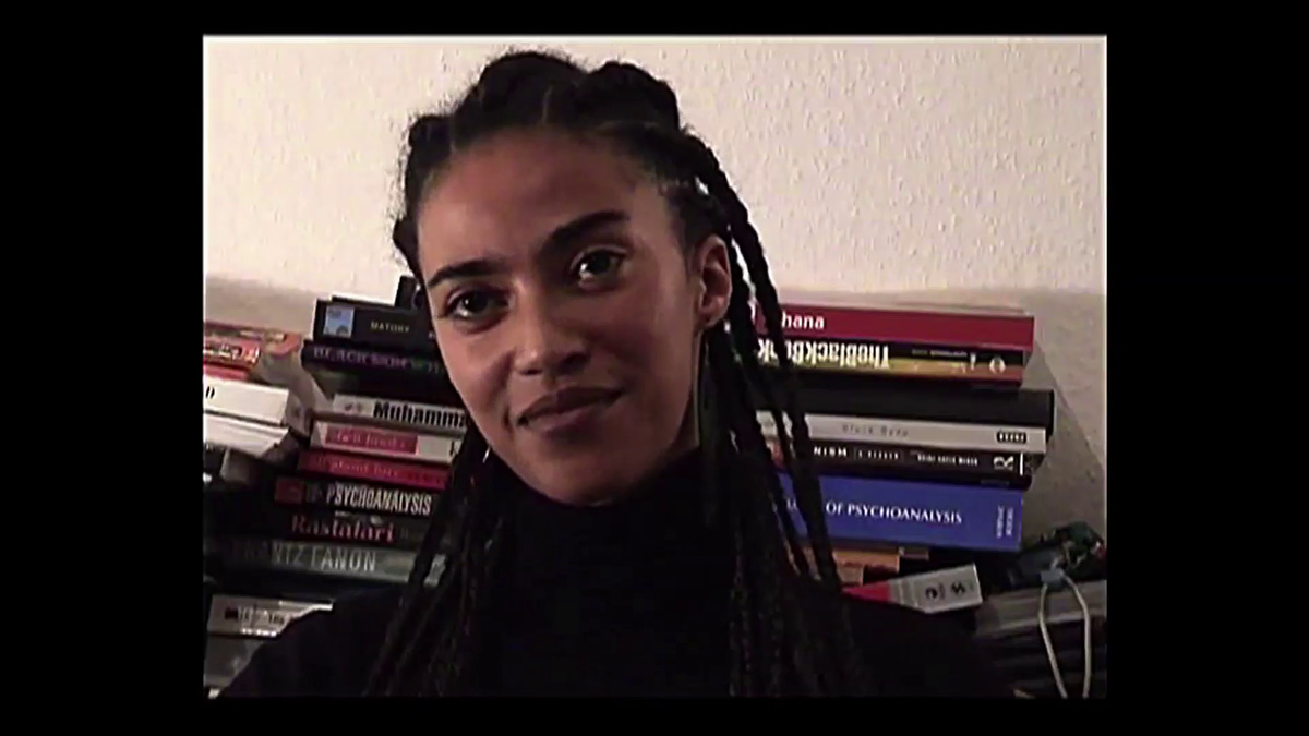 A still from an interview, the subject is smiling and wearing a black turtle neck, they are in front of piles of books