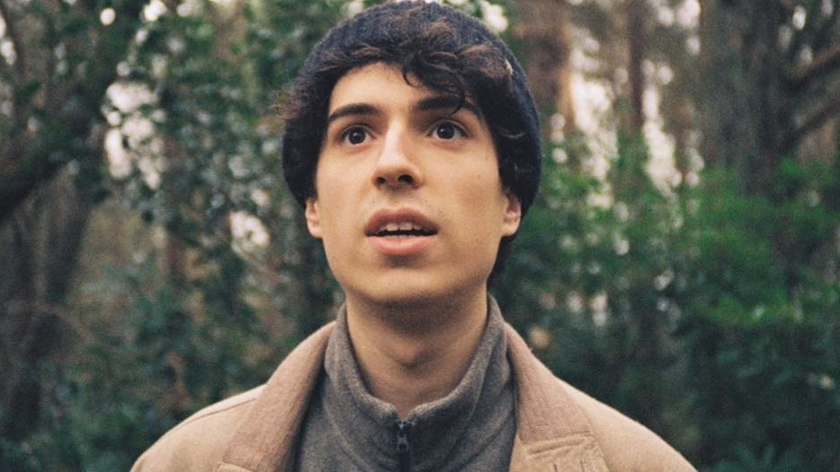 A young man wearing a wooly hat, fleece, and sports jacket stood in front of a wooded area.