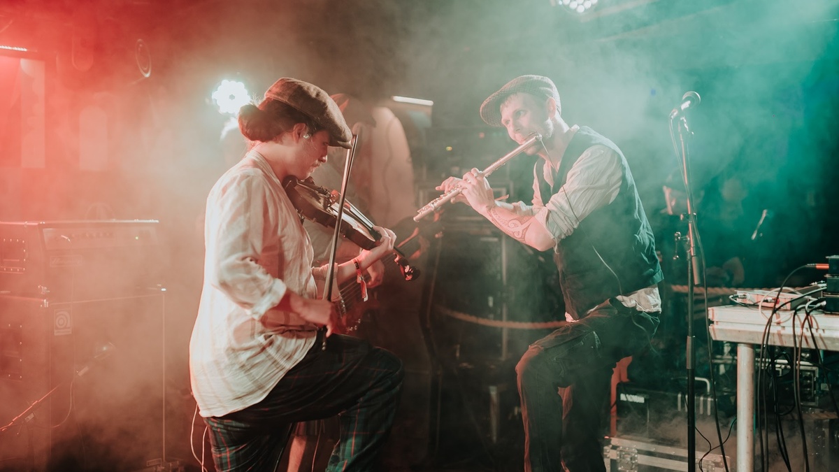A duelling fiddler and flautist playing against each other on a smokey stage surrounded by various musical equipment.