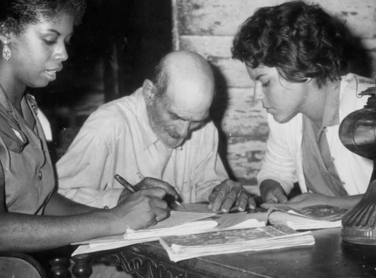 A black and white image of 2 women teaching an elderly man, the man is writing whilst one of the teachers watches.