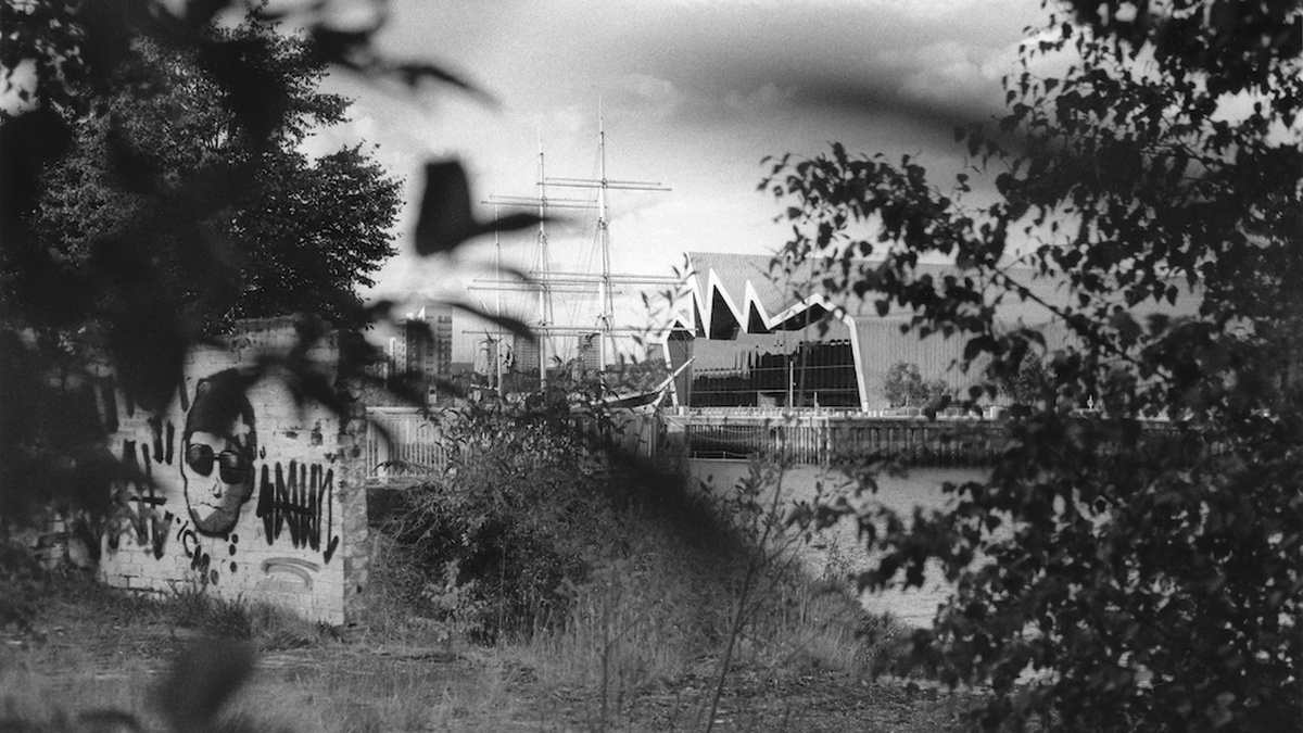 A black and white image of Glasgow's Transport Museum and Tall Ship taken from afar, obscured by leaves and branches.