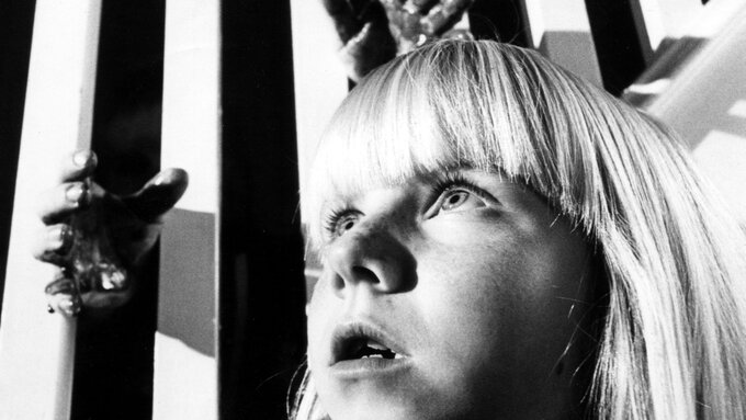 A B&W still, a woman in the foreground with white banisters behind her, a pair of hands reach out from the darkness.