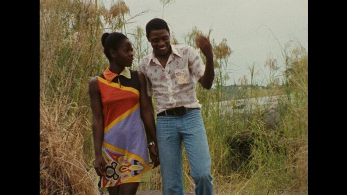 2 people, 1 in a colourful dress and the other in jeans and a shirt, walk along the tall grass and the edges of a lake.