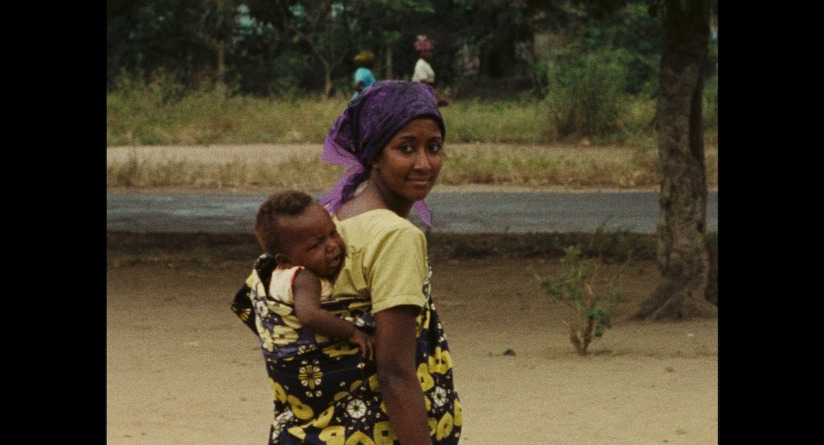 A woman with a baby in a wrap of fabric on her back looks back towards the viewer, she wears yellow and purple clothes.