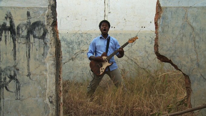 A man playing electric guitar faces the camera. He stands in dry grass and there are destroyed walls around him.