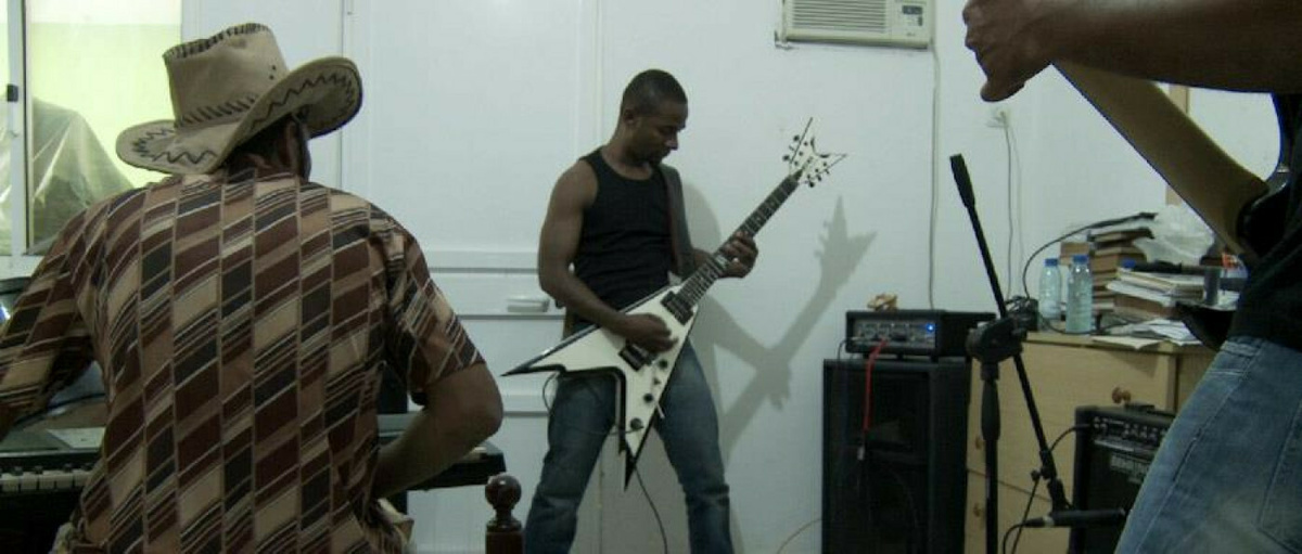 Three band members playing two electric guitars and a keyboard are rehearsing in a room.