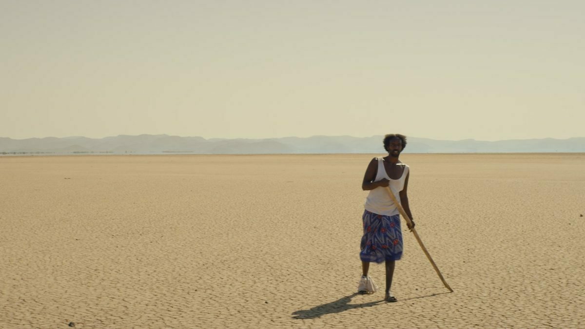 A lone man stands in a wide flat desert, he has one bandaged foot and is using a stick to walk.