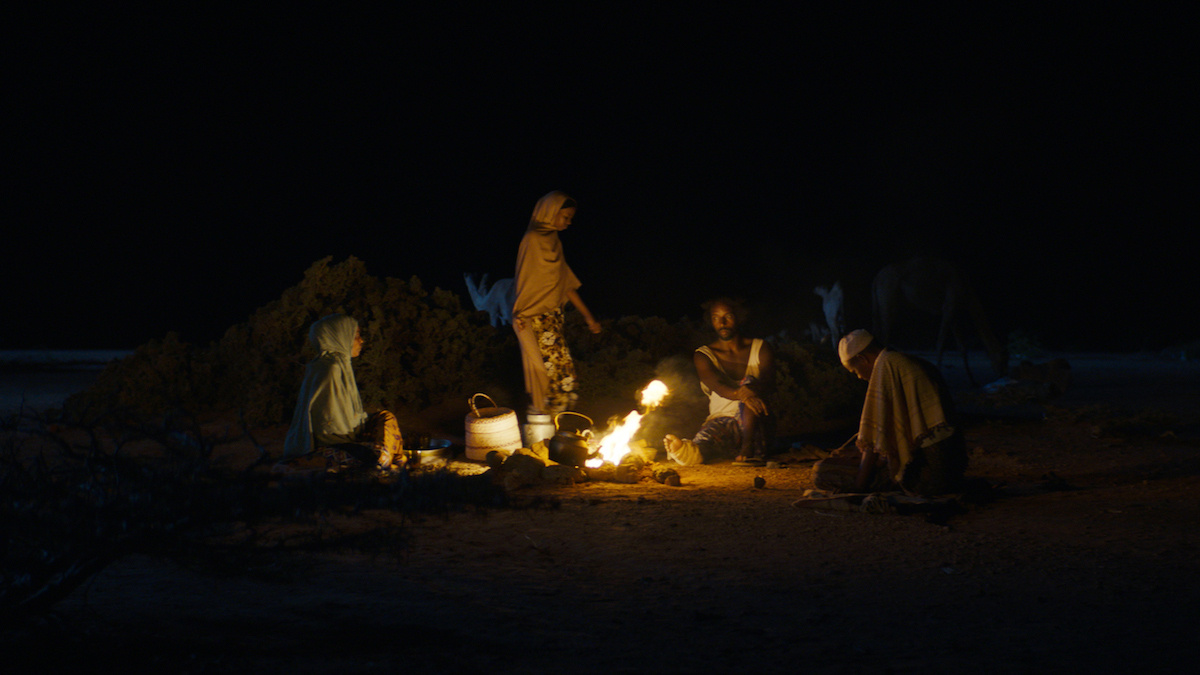 4 figure around a campfire 1 stands whilst the other 3 sit, there's a kettle on the fire, and figures in the background.