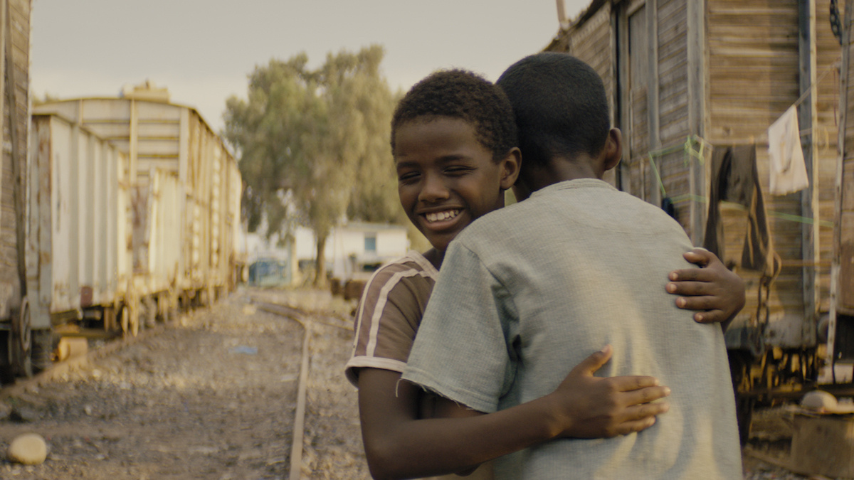 Two young children hug each other, the one facing us is smiling. They are situated in a train depot.