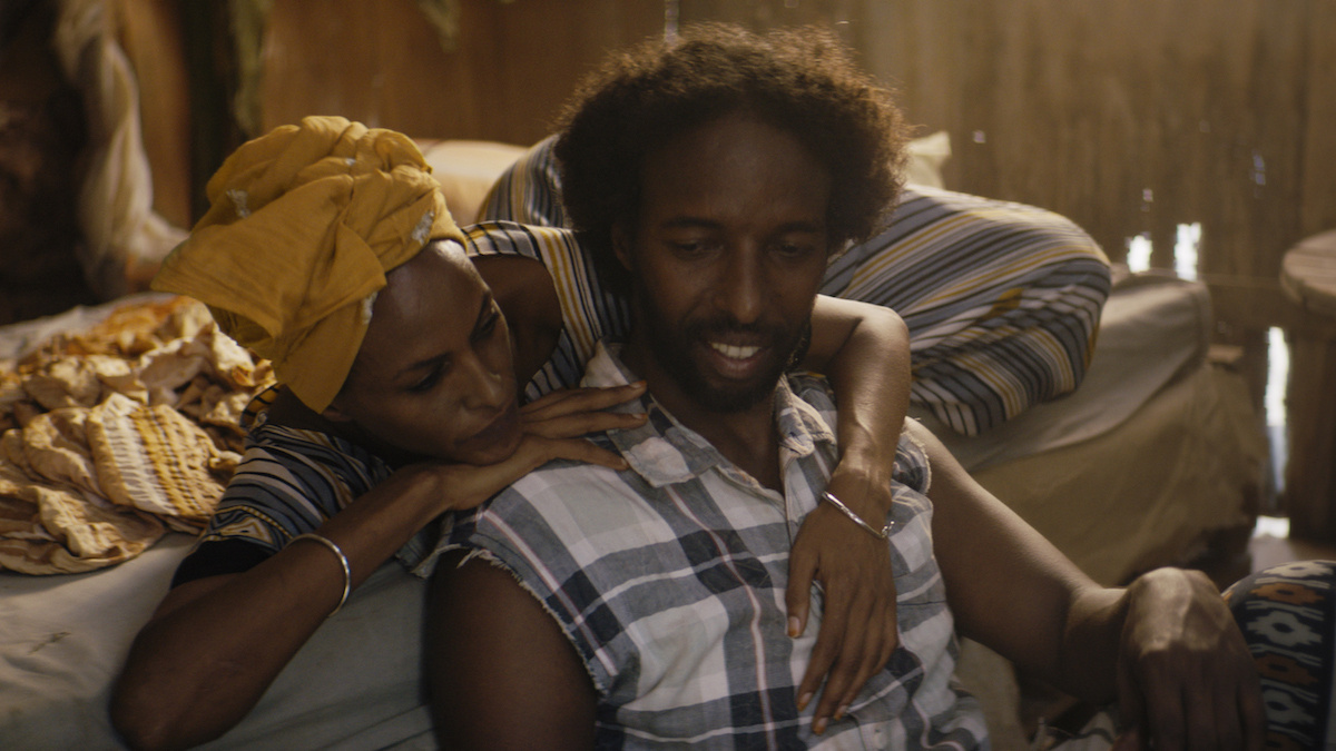 A man in a checkered shirt sits against a bed where a woman lays with her arms wrapped around him, they look peaceful.