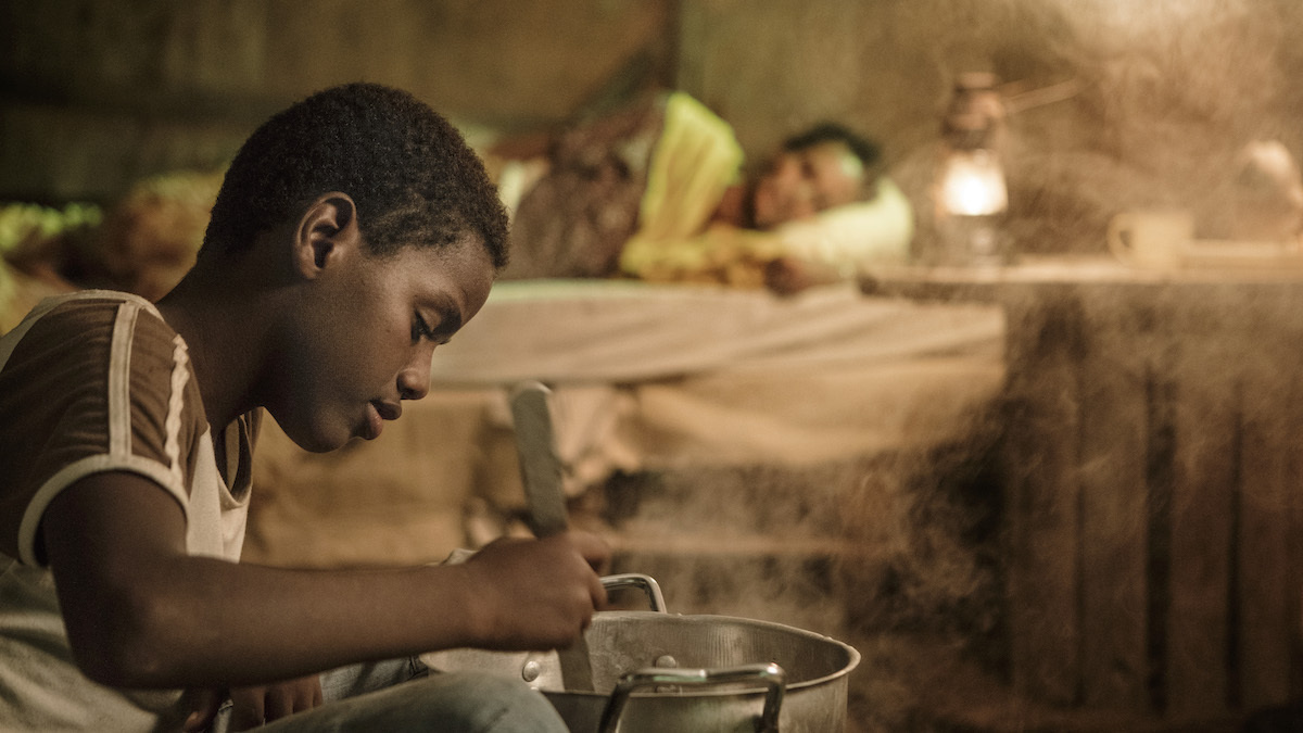 In the foreground a young boy cooks something in a pot, whilst an elderly person lays in a bed behind him.