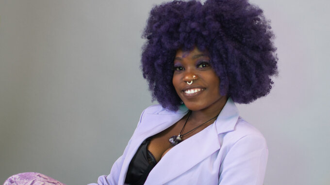 EYVE sits reclined on a chair, smiling at the camera. She has purple natural hair.