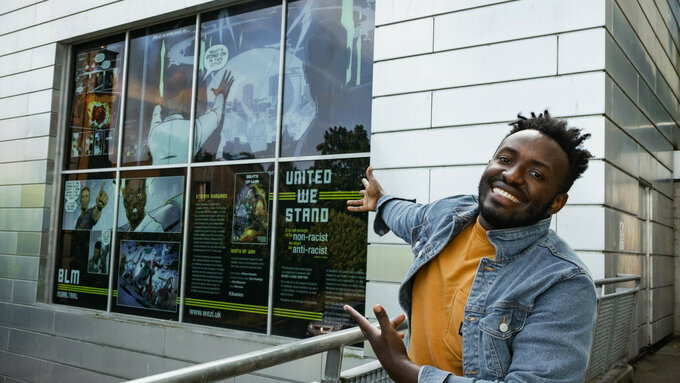 Etienne Kubwabo gesturing at a large display of his comics in a window. He's smiling brightly, wearing an denim jacket.
