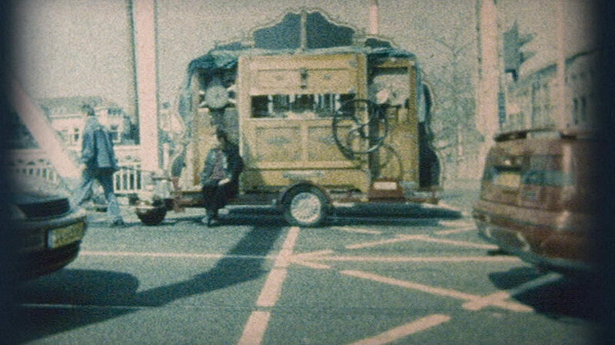 A grainy still image of a street scene. In the centre of the image a person sits on a parked, unusual yellow vehicle.