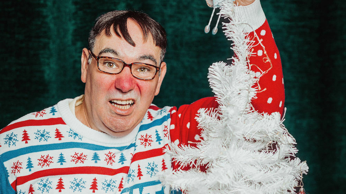 A fat bespectacled red-nosed man in a Christmas jumper grimaces putting a fairy decoration on top of Christmas tree.