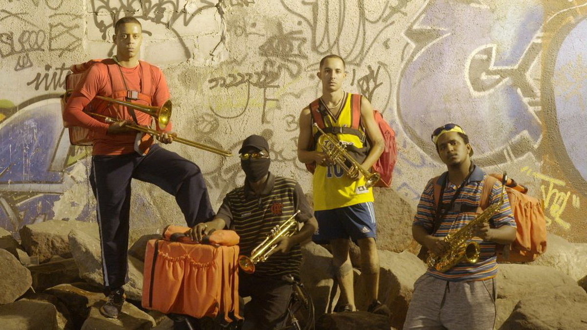 Four male food delivery riders holding trumpets and saxophones are posing against a graffiti-covered wall.