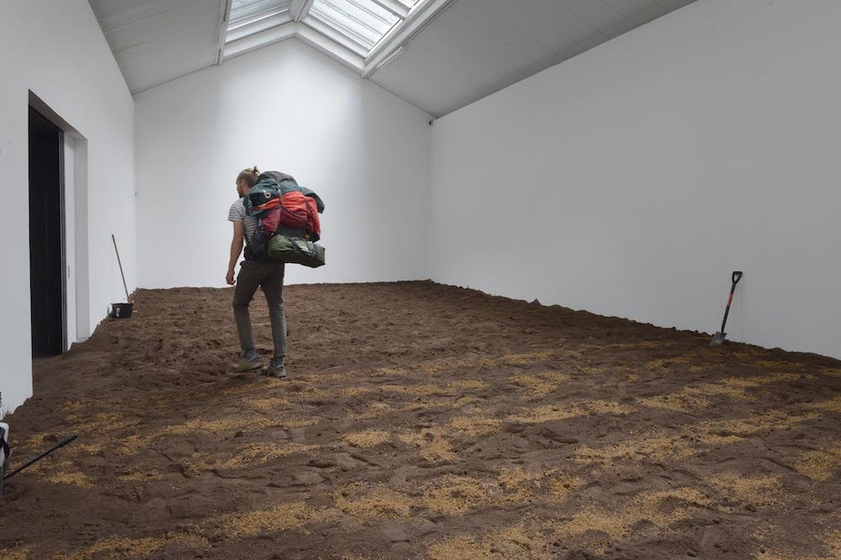 A room full of soil with a person wearing a heavy hiking backpack striding across it.