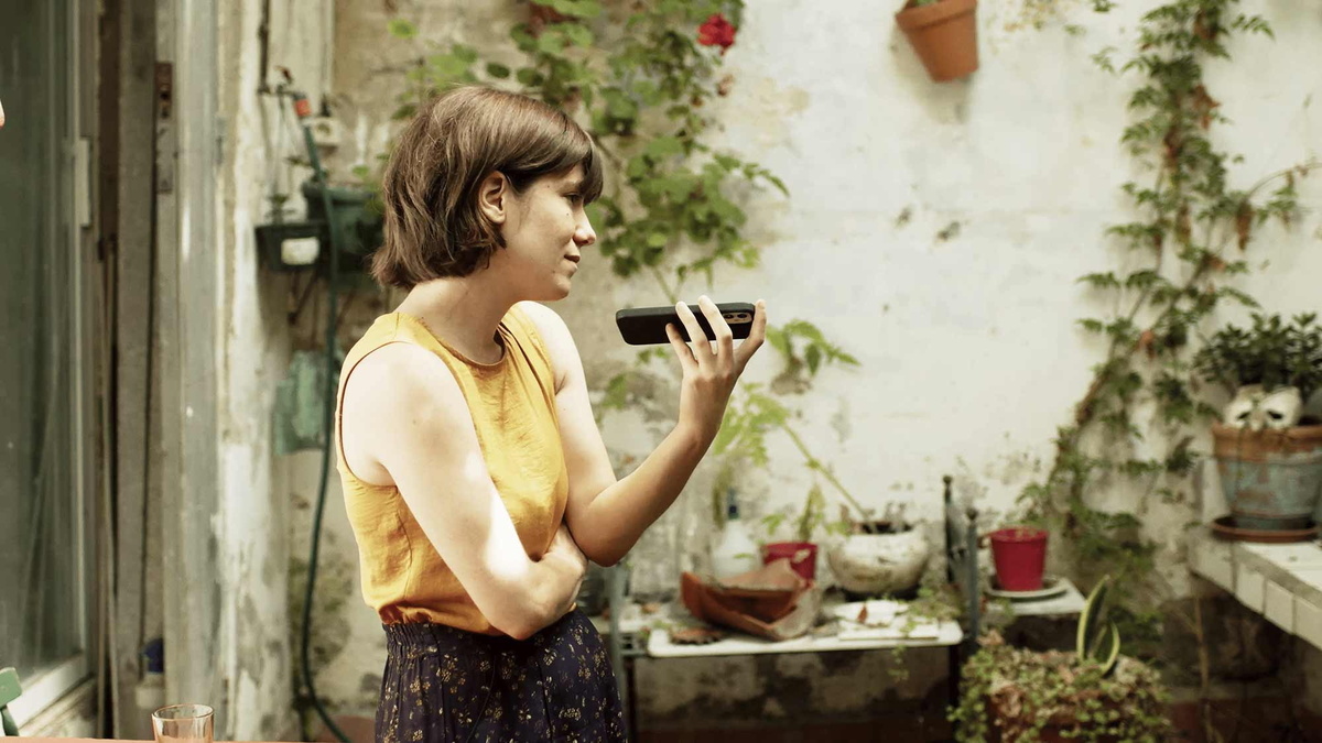 A young woman recording a voice note on her phone while standing in a patio full of house plants.