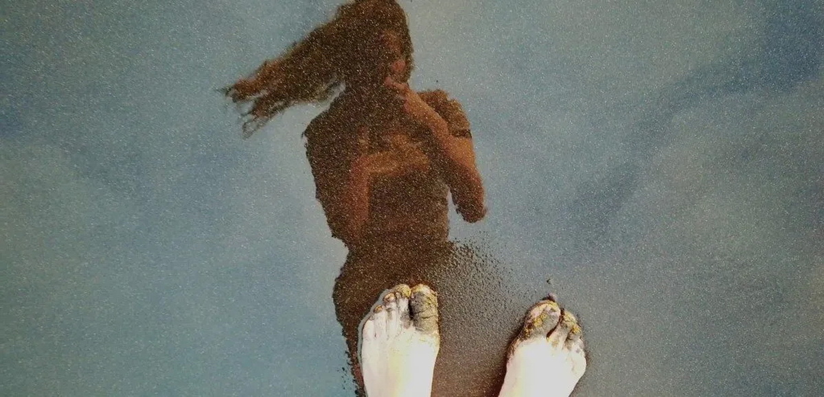 A woman is taking a photo of her bare feet and her reflection in the wet sand.