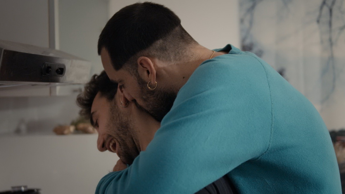 Two men are pictured standing in the kitchen. One of them is wearing a blue pullover shirt and is hugging the other man.