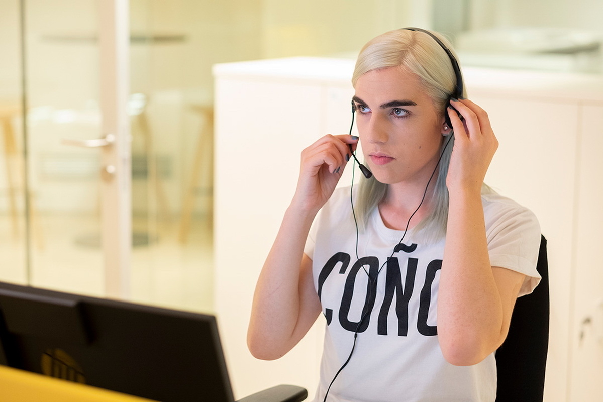 ain character Raphi, a trans woman now with blonde hair, is pictured in a call centre with a headset on.