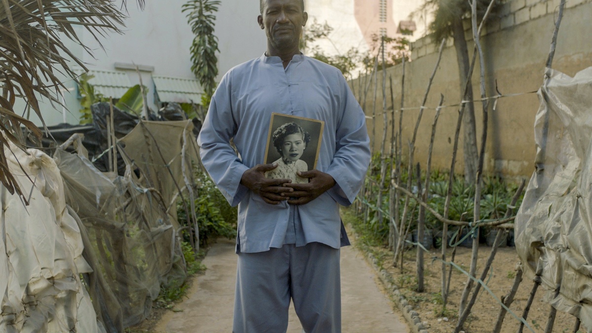 A dark skinned man stands in the centre of a leafy path. He is holding a black and white framed portrait of a woman.