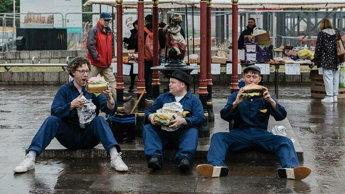 Three workers in blue boiler suits sitting on a step in front of a market in Govan eating comically big sandwiches.